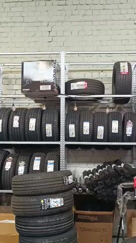 The video shows an interior view of a storage or retail space, likely a tire shop or auto parts store. Metal shelving units are filled with various items. On the shelves, there are stacks of tires, boxes of various sizes, and some miscellaneous items such as car oil containers, tool boxes, and black plastic parts. Some of the boxes have branding visible, including "YATO" and "KRAFT&DELE." In the lower part of the frame, a stack of tires is leaning against a shelf, and in the foreground, the corner of a black tire is visible. The walls appear to be made of brick. The lighting suggests an indoor environment. No people are visible in the video.