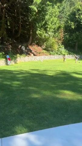 Two children are playing soccer on a grassy lawn in front of a dense forest. The child on the left is wearing a white t-shirt and shorts, and the child on the right is wearing a white t-shirt and shorts. The child on the left kicks the ball towards the child on the right. A person in a red shirt is visible in the background, standing near a stone wall. The sun is shining, and there are shadows on the grass.