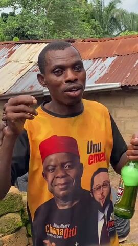 The video shows a Black man in a yellow t-shirt with an image of a politician on it. He is holding a green bottle of beer in his right hand and a small object in his left hand, which he is about to place in his mouth. The man is looking into the distance and appears to be speaking. In the background, there are other people and a corrugated iron roof. The environment appears to be outdoors.