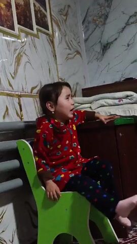 A young girl is sitting on a green child's chair in what appears to be a bedroom. She is wearing a red long-sleeved shirt with a pattern of hearts and other shapes, and black leggings with colorful stars. The girl has her hair pulled back and is wearing small earrings. She is looking up and to the right, and at one point appears to be crying or upset. Her hand is resting on a dark wooden cabinet or nightstand next to a bed that is partially visible in the background. The room has wallpaper with a marble-like design in shades of white, gray, and gold. There are three framed pictures hanging on the wall above and to the left of the chair. A gray metal structure is also visible to the left of the chair.