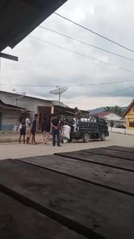 A group of young people stand on a paved road next to a small black truck. The truck is loaded with large white sacks. One person is standing in the truck bed, holding onto a sack. Another person appears to be directing them. The scene takes place in a rural or semi-rural area with houses and satellite dishes visible in the background. The sky is overcast. The view is framed by wooden planks in the foreground.