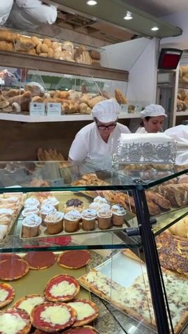 The video shows a bakery. Two women in white baker's hats and uniforms are behind a counter. The woman in the foreground is wearing glasses and is looking to her right. She is in front of a display case filled with baked goods, including small cakes topped with powdered sugar, pizzas, and other pastries. The woman in the background is facing away from the camera, reaching for something on a shelf behind her. Shelves stocked with bread are visible in the background. The environment appears to be a well-lit retail space with glass display cases and mirrors.