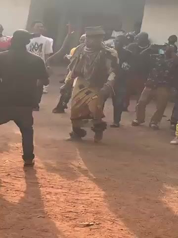 A crowd of people have gathered outdoors on a dirt ground outside a building. A person dressed in traditional clothing and carrying a stick is dancing in the middle of the group. Some members of the crowd are also dancing or swaying to the music. Many people are wearing casual clothing like jeans, t-shirts, and sneakers. The scene appears to be taking place during the daytime.