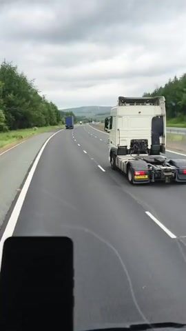 The video shows a view from inside a vehicle driving on a highway. The main subject is a white semi-truck driving in the same direction, ahead of the camera vehicle. Another blue semi-truck is visible further ahead on the highway. The highway is paved and has white lane markings. On either side of the highway, there are trees and grassy hills. The sky is overcast with clouds, suggesting a possibly gloomy or rainy day. In the foreground, a dark object, likely a smartphone, is partially visible at the bottom of the frame, indicating the video is being recorded from a handheld device. The license plate of the white semi-truck is partially visible and appears to read "CY23 FWB". The location is identified as Biggar, United Kingdom. The overall scene conveys a sense of travel on a highway under cloudy weather.