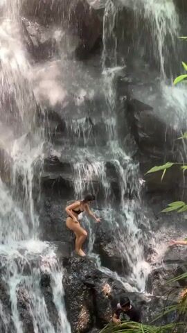 A daytime video captures several people at a natural waterfall. A woman in a black bikini stands on the wet, dark rocks of the waterfall, looking down as water cascades around her. Further down and to the right, another person wearing a straw hat and swimwear is partially visible. At the bottom of the frame, a man in a dark shirt and cap is crouched, seemingly examining something on the ground. Lush green foliage frames the right side of the scene, indicating a jungle or forested environment. The activity appears to be recreational, with people exploring and enjoying the waterfall. The setting is bright and natural.