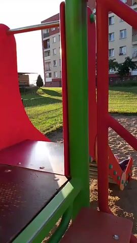 A young child with blonde hair, wearing a pink shirt, is sitting on a red playground slide. The child is looking towards the camera. The slide is made of red plastic and has wooden supports underneath. The playground has a sand surface. In the background, there is a green fence, a grassy area, and a multi-story apartment building. The sun is shining brightly, creating lens flares in the video. The camera is moving, as indicated by the slight blur and changing perspective.