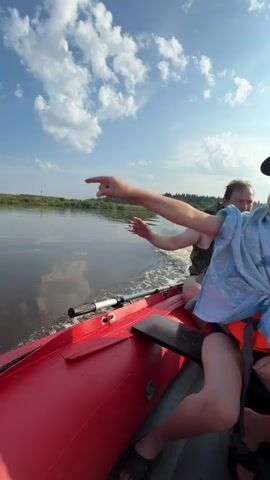 A group of people are on a boat ride. The boat is red and appears to be an inflatable raft. There are at least three people visible, with a young boy in the foreground wearing a green hat and a blue towel draped over his shoulders. He is also wearing an orange life vest. Behind him, an adult male wearing a life vest is operating the boat. Further back, another person is visible wearing a life vest. The boat is moving through water, leaving a wake. The environment appears to be a river or large body of water, with a shoreline of trees and vegetation in the background. The sky is blue with scattered white clouds, and the sun is shining. The camera angle is low and seems to be handheld, looking forward and slightly to the side.