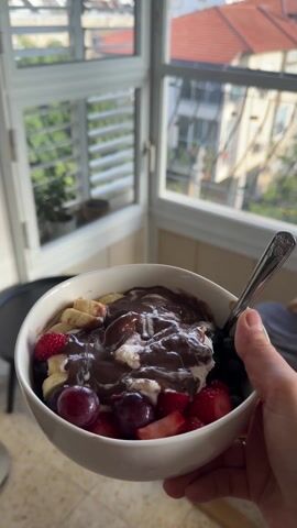 A hand holds a white bowl filled with fruit, ice cream, and chocolate sauce. The fruit includes blueberries, sliced bananas, halved strawberries, and cherries. A silver spoon rests in the bowl. The bowl is held in front of a window. The background shows a green and red rooftop across the street.
