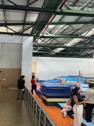 The video shows an indoor gymnasium with various gymnastics equipment, including mats and a balance beam. In the foreground, two young women stand near a railing. To their right, a man in a red shirt and dark pants appears to be coaching or instructing a young gymnast. The gymnast, wearing a black leotard, is in the process of performing a handstand on a stack of blue mats. She then transitions out of the handstand and lands on the mats. The gym has a high ceiling with exposed metal beams and pipes. Several other people, including children, are visible in the background, observing the gymnastics activities. The overall atmosphere seems to be one of training or practice.
