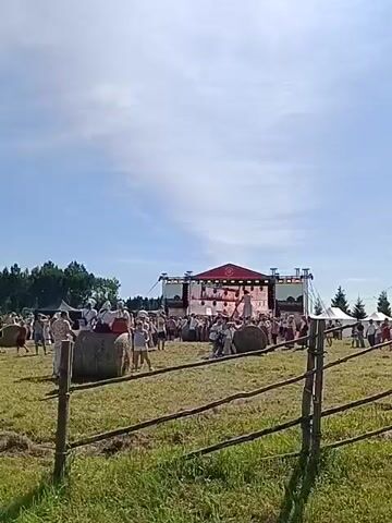 A wide shot captures an outdoor music festival. In the foreground, a rustic wooden fence made of branches divides a grassy field from the main event area. Several large hay bales are scattered across the field, serving as impromptu seating for some attendees. In the mid-ground, a large stage is set up with a screen displaying images and a red-roofed canopy. A crowd of people, dressed in casual summer attire and some in what appears to be traditional clothing, are gathered in front of the stage, watching a performance. Some individuals are seated on the hay bales, while others stand and dance. To the right of the stage, a white tent is visible, possibly for vendors or shelter. The background features a treeline and a bright blue sky with scattered clouds, suggesting a sunny day. The overall atmosphere appears festive and relaxed.