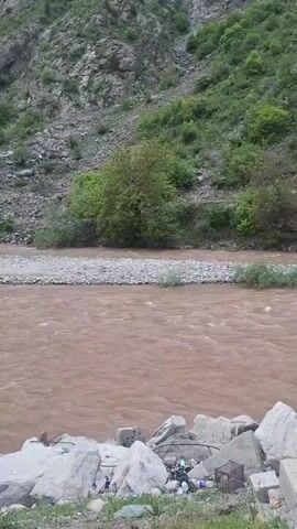The video shows a wide river flowing between rocky banks, with a hillside visible in the background. The water is brown and muddy-looking, indicating a high sediment content or recent heavy rainfall. The rocky banks are lined with gray boulders and some vegetation. The hillside in the background is covered with sparse greenery and rocky outcroppings. There is also trash at the rocky bank.