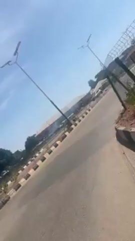 The video, taken from what seems to be a moving vehicle (possibly a motorcycle or auto rickshaw, judging from the handlebars in view), depicts a roadside scene on a sunny day with a bright blue sky. The road is asphalt with black and white-striped concrete lane dividers. A fenced-off area runs parallel to the road, containing reddish-brown dirt and sparse vegetation. In the background is a gas station, as well as electricity poles. The camera angle is unstable due to the moving vehicle.