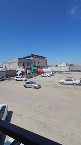 The video shows a bright, sunny day with a clear blue sky and some wispy clouds. The scene appears to be an outdoor lot, possibly a truck stop or a depot, with a dusty, unpaved surface. In the foreground, a dark railing or ledge is visible, suggesting the video is filmed from an elevated position.

A variety of vehicles are present. To the left, a white convertible is parked near a building. Behind it, a large white truck with its cab tilted upwards is partially visible. Next to the tilted truck is a green truck, and to its right, an orange and white tanker truck. Several smaller cars, including a silver sedan in the center of the frame, are parked or driving in the lot. Further back, numerous white semi-trailers are lined up. A white older model sedan is parked on the right side of the frame, and a gray sedan is parked next to it. Another large white truck is on the far right.

The building in the background has a red sign with white lettering. There are no people clearly visible in the video. The overall impression is of a busy industrial or transportation hub on a warm, clear day.