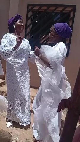 The video shows three women dancing. They are all dressed in white robes and purple head coverings. The woman in the center has white lace on her dress. The other two have flowing fabric sleeves. The ground they are on looks like sand. A building with a barred window is behind them. Other people are nearby.