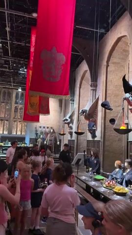 The video shows a large hall with high stone walls and arched windows. Red banners with golden crests hang from the ceiling. Several large, wing-like sculptures are mounted on the walls, each holding a flaming torch and a witch's hat. In the foreground, a long table is set with plates of food and drinks. Several mannequins dressed in Hogwarts robes are seated at the table. A group of people, mostly women and girls, are standing on the left side of the frame, looking towards the table and the hall. One person in the center appears to be speaking. Hats are seen floating in the air above the table. The scene appears to be a reenactment or exhibit of a scene from the Harry Potter movies.