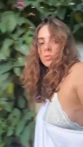 A woman with wavy brown hair, wearing a white top and a white robe over her shoulders, walks onto a patio. She is holding a camera. The patio has potted plants and overlooks a green lawn and a gazebo, with a view of rolling hills in the distance under a blue sky with white clouds. The camera then tilts up to focus on the sky.