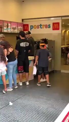Here's a description of the video:

Outside a McDonald's, a group of children are gathered around a small promotional table. A McDonald's employee with dyed orange streaks in his hair stands behind the table, interacting with the children. The table has a sign that says "Adivina el Orden!!" which translates to "Guess the Order!!" There are cutouts of McDonald's food items like fries, a drink, an ice cream cone, and a burger on the table. Other customers are visible at the McDonald's dessert counter in the background. The scene is bright and sunny.
