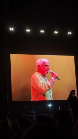 An audience is watching a live performance on a large screen. The screen shows a man with white hair wearing a light green shirt, holding a microphone and talking into it. The scene then changes to a wider shot on the screen, revealing that the man is on stage with another person playing an accordion. The stage is in front of a large crowd of people. The lighting on stage is red. The audience members are recording the performance with their phones.