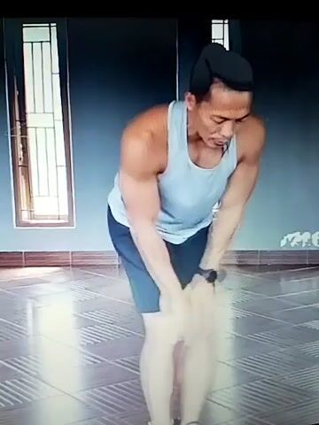 A man in a white tank top and dark shorts is performing a physical exercise. He is bending at the waist with his knees slightly bent and his hands placed on his knees. His head is lowered as he looks down. The background appears to be an indoor space with tiled flooring and a window with vertical blinds on the left side of the frame. A microphone is attached to his ear. The top of the screen displays text in a language other than English, potentially indicating a title or description of the video. The man's muscular physique is visible.