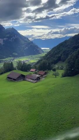 The video shows a high-angle drone shot of a picturesque mountain valley. In the foreground, a vibrant green, gently sloping meadow is dotted with a cluster of rustic wooden houses with dark roofs. The houses are nestled close together, with a larger structure on the left and smaller ones to its right. A few trees are scattered across the meadow, one particularly noticeable on the right side of the houses.

The middle ground of the valley floor is a tapestry of green fields and darker patches of trees, suggesting a small village or hamlet. A winding road or path is visible, disappearing into the greenery.

In the background, towering, rugged mountains rise steeply on both sides of the valley. The left mountain range is imposing and rocky, with some lighter patches that could be snow or rock faces. The right side is densely covered with dark green forest.

At the far end of the valley, a body of water, possibly a lake, is visible in the distance, reflecting the sky. The sky itself is dramatic, with large, fluffy white clouds interspersed with patches of bright blue. Sunlight streams through gaps in the clouds, casting distinct patterns of light and shadow across the landscape. The overall impression is one of serene, natural beauty.