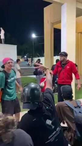 The video shows a group of people gathered outside at night, possibly for an event or gathering.  Several individuals are standing, while others are seated. A person in a red jacket stands near the edge of the crowd. Another person, wearing a green shirt, a pink hat, and carrying a backpack, gestures towards the person in the red jacket while speaking. In the foreground, a person with a motorcycle helmet sits with their back to the camera. Other individuals are scattered throughout the scene, and in the background, there is a statue on a pedestal. The surroundings appear to be a park or public space with buildings and lights visible.
