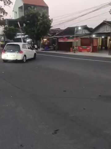 The video shows a street scene in what appears to be Southeast Asia, likely Indonesia. The road is asphalt and has a pedestrian crossing. On the left side of the road, there is a white car parked, and a tree. Further down the street, there are several small shops and buildings. One building has a large banner with text and images. Another building appears to be a shop with goods displayed outside. There are several motorcycles parked in front of the shops, and a few people are visible, though their faces are blurred due to the low resolution and movement. On the right side of the road, there are more shops, some with awnings and lights. A black pickup truck is seen on the road. The sky is overcast, suggesting it might be dusk or dawn. The overall impression is a bustling street in a residential or commercial area.