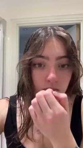 Here's a description of the video:
The video features a woman with brown hair and light eyes, wearing a black tank top, in front of a bathroom. Her hair is wet and loose. She has a nose piercing and is wearing a necklace. The woman is putting on mascara. The bathroom behind her has white tiles and a door frame with a door ajar.