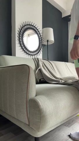 A woman with long brown hair pulled back in a ponytail is shown arranging a throw blanket on a light green couch. The couch is against a wall that is painted white on top and dark green on the bottom. A round mirror with decorative black spokes hangs on the wall above the couch. A floor lamp stands to the right of the mirror. The woman is wearing a white t-shirt, light green shorts, and a watch. The floor is made of light wood planks.