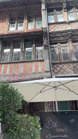 The video shows an exterior view of buildings with timbered facades. In the foreground, there is a leafy green tree and several white umbrellas, suggesting an outdoor seating area for a cafe or restaurant. A sign with the word "Motel" in pink lettering is visible on one of the buildings. People are seen walking in the background and sitting at tables. The camera pans to show more of the street with similar architecture and more outdoor seating. The scene appears to be a lively street with businesses operating.