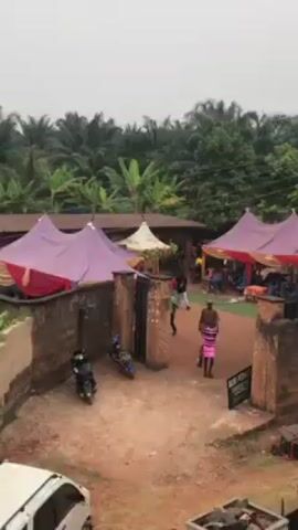 The video depicts an outdoor scene, seemingly in a rural or semi-rural area, where a gathering or event is taking place. Several tents with pink and yellow canopies are set up on a dirt ground, suggesting an organized function. People are seen walking around and sitting under the tents, which indicates a social event is in progress. The background features dense vegetation, including palm trees, suggesting a tropical or subtropical climate. A stone wall or structure is visible in the foreground, partially obscuring the view. The overall atmosphere suggests a community event or celebration in a natural setting.