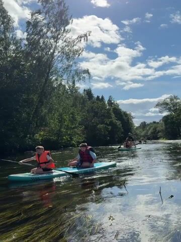 A group of people are paddleboarding down a river. The water is murky and green with aquatic plants. The banks of the river are lined with trees and lush green foliage. The sky is blue with scattered white clouds. The people are wearing life vests and appear to be enjoying the activity.