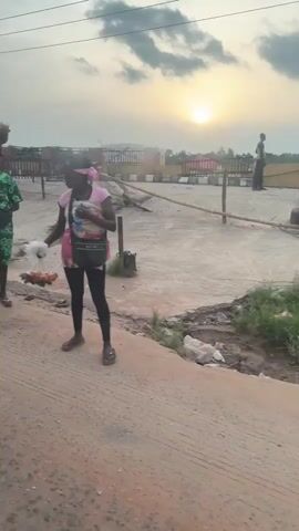 The video shows a dirt road with a deteriorating concrete surface, flanked by a fenced area with sparse vegetation and a setting sun in the distance. Two individuals are visible: a person in green carrying a basket on their head and another person in a pink shirt holding a bundle of what appears to be eggs. The camera appears to be inside a vehicle.