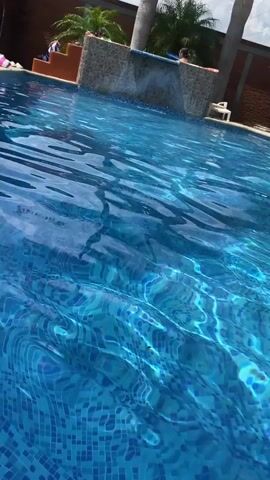 The video shows a person swimming in a clear blue swimming pool. The pool has a mosaic tile bottom and is surrounded by palm trees and a brick wall. The water is rippling, creating a shimmering effect. A small waterfall feature is visible at one end of the pool. The sky is blue with scattered clouds. There is a white lawn chair near the wall and other people visible on the upper level by the waterfall feature. An electrical tower is also seen in the background. The camera pans slightly upwards.
