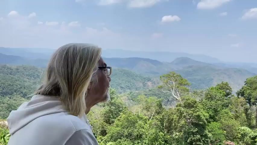 A man with long white hair, a beard, and glasses stands against a background of rolling green hills. The sky is light blue with some white clouds. He is wearing a white hooded sweatshirt. He looks to the left and then turns toward the camera, gives a thumbs-up, and then looks away. The camera then pans away from him, focusing on the mountains.