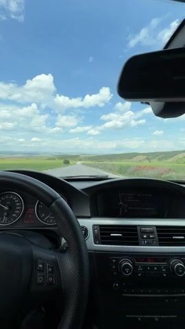 The video shows a first-person perspective from inside a moving car. The car is driving on a paved road through a rural area. The sky is blue with scattered white clouds. The landscape consists of green fields and some rolling hills in the distance. The car's dashboard and steering wheel are visible, showing the car is a BMW. The car is traveling on a straight road, and there are no other vehicles immediately visible. The side mirror reflects the road and landscape.