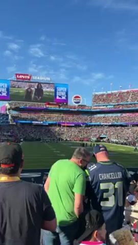 The video shows a crowded football stadium under a blue sky. People are in the stands, and many are wearing team jerseys. A man in a green shirt and a man in a Seattle Seahawks jersey are standing in the foreground. A person wearing a black coat and beanie walks by carrying a colorful snack. The camera pans up to show the stadium's upper levels and the crowd.