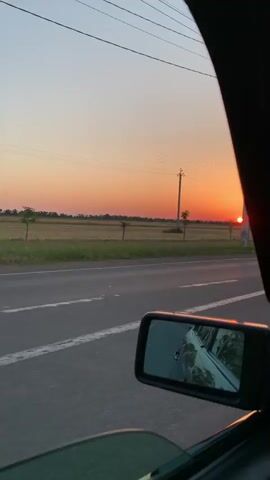 The video shows a view from inside a moving car, looking out the side window. The sun is setting on the horizon, casting a warm orange glow across the sky and fields. There are telephone poles and power lines visible against the sky. A flock of birds is flying in the distance. The car's side mirror reflects the road and the surrounding landscape. The road is asphalt and has white lane markings. The fields are green and appear to be agricultural land. The overall atmosphere is serene and peaceful, with the warm colors of the sunset creating a sense of tranquility.