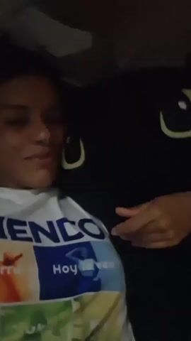 A woman with dark, curly hair and medium-toned skin is lying down, with a large black object, possibly a pillow, next to her. She is wearing a white t-shirt with blue and colorful graphics. The graphics on her shirt appear to be a logo with Spanish text. The lighting is dim. A hand, with medium-toned skin, is visible on the right side of the frame, pointing towards the woman's shirt. The woman smiles at the camera in the initial frames, then her expression becomes more neutral.