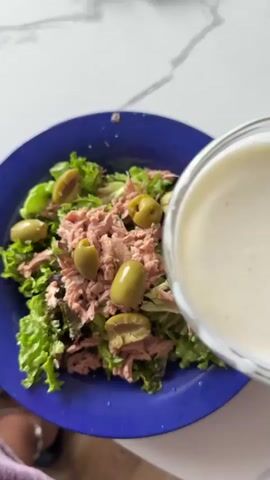 A blue plate of tuna salad with lettuce and olives sits on a white countertop. A glass bowl of white dressing is being poured over the salad. A hand with pink nail polish is holding the bowl. A fork is visible to the right of the plate. The salad consists of shredded lettuce, canned tuna, and green olives. The dressing appears to be creamy and thick.