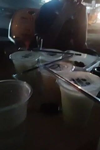 Here's a concise description of the video:

The video appears to be shot outdoors at night, likely at a street food vendor. A person is seen sitting behind a table. There are several plastic cups filled with a light-colored drink on the table, labeled with the word "DRINK." Straws are sticking out of the cups. There is also food in plastic containers on the table. The lighting is poor, making it difficult to discern specific details. In the background, there are blurry lights of passing vehicles.