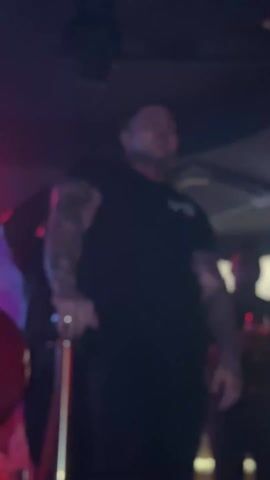 The video shows a man standing in a nightclub, gesturing a heart shape with his hands. He is wearing a black t-shirt with a logo on the front, dark pants, and has tattoos on his arms. The lighting in the club is dark, with a red light shining down from above. There are other people in the background, including a woman with a red sequined outfit and a man holding a wine glass. A gold stanchion and red velvet rope is visible to the man's left.