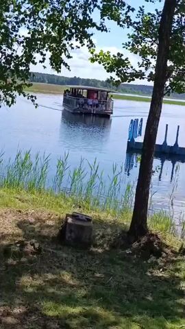 The video shows a scenic view of a lake on a sunny day with scattered clouds. A small boat with people on deck is moving away from the shore. In the foreground, there's a wooden pier extending into the water, with several individuals standing on it. The pier appears to be made of modular plastic floating platforms. A tree stands on the grassy bank near the pier. The water is calm, reflecting the sky and surrounding greenery. The overall atmosphere is peaceful and suggests a recreational setting, possibly a tourist attraction or a local gathering place.