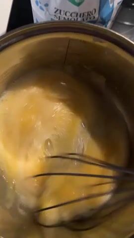 The video shows a top-down shot into a stainless steel pot containing a yellow liquid being vigorously whisked. The liquid appears to be a mixture of eggs and possibly sugar, given a bag of "Zucchero Semolato" (Italian for granulated sugar) is visible in the background. The whisk moves rapidly, creating small bubbles on the surface of the liquid. The pot is on a dark countertop. The camera remains mostly still, focused on the mixing process.