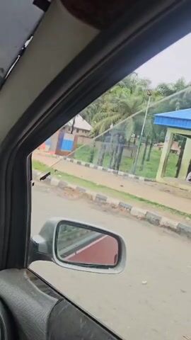 The video is shot from inside a car, looking out the passenger side window. The camera pans slowly across the scene. Outside the car, palm trees are visible along a road. There are piles of sacks or bags stacked along the roadside, some covered with tarps. Further down the road, there are several vehicles, including trucks and what appear to be motorcycles. People are also visible on the side of the road. The road is paved and bordered by a striped curb. The overall impression is that of a busy roadside scene in a tropical or subtropical location.