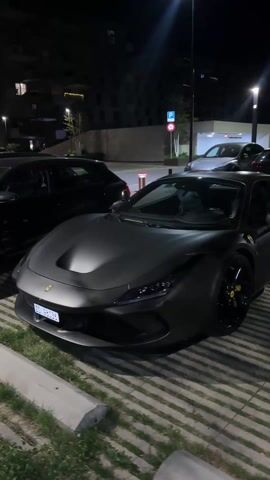 The video shows a matte black Ferrari parked on a striped, paved surface at night. The car has black wheels and yellow brake calipers, with a Ferrari logo visible on the front quarter panel. A black car is parked to the left, and other cars are visible in the background. Buildings are visible behind the cars, lit by streetlights. A parking sign is visible near the background.