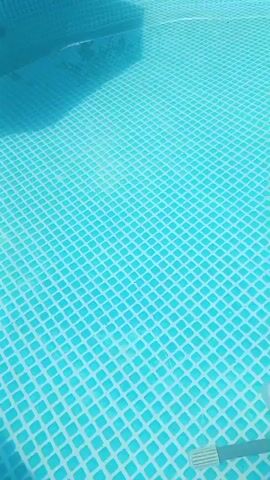 The video shows a close-up of an above-ground swimming pool. The water is a bright blue color and the bottom of the pool has a white tiled pattern. In the first few seconds, there is a pool skimmer or vacuum being used to clean the pool. The skimmer has a black hose attached to it. The water is disturbed by the cleaning device, creating ripples and bubbles. The video then pans up to show the edge of the pool and then shows a wider shot of the pool with an umbrella providing shade. Lush green foliage is visible in the background, along with a bright blue sky.