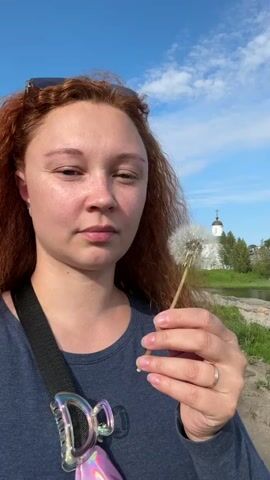 A young woman with red hair and closed eyes blows on a dandelion seed head. She holds the dandelion in her right hand, and it is positioned in front of her face. The background is out of focus but appears to be a rural outdoor setting with a blue sky, clouds, trees, a body of water, and a white building with a dark dome, possibly a church. The woman is wearing a navy blue shirt, and a strap from a bag or camera is visible across her shoulder. A ring is on her right ring finger. As the video progresses, the dandelion seeds begin to disperse. The woman then opens her eyes and smiles at the camera.