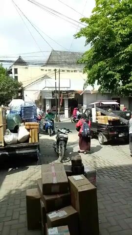 The video shows a street scene with several vehicles and people. In the foreground, there are stacks of cardboard boxes. To the left, a truck is loaded with goods, including what appears to be furniture wrapped in plastic and rolls of material. In the middle ground, a black pickup truck is also loaded with items. Two men are near the pickup truck, one appears to be leaning into the bed of the truck. A woman in a red shirt and dark skirt is walking away from the camera towards the pickup truck. A motorcycle is parked in front of the woman. Another motorcycle, ridden by a person in a green shirt, is moving on the street. In the background, there are buildings with awnings and trees lining the street. The sky is visible and appears to be overcast.