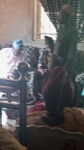 Okay, here's a description of the video:

The video shows a small gathering of people in what appears to be a living room. There are at least four individuals visible. One man is seated on a sofa wearing a white robe and a light blue cap. He is sitting near a small wooden table with two bottles on it and appears to be examining something in his hands. Another man, seated next to him on the sofa, wears a patterned top and a black hat. A woman is seated on the floor near the sofa. The environment seems to be dimly lit and the interior design is somewhat dated. The room contains sofas, a window, and some decorative elements.