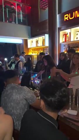 The video shows a lively scene inside a bar or nightclub. In the foreground, several people are gathered around a bar counter, with one person appearing to be a DJ. The DJ is a woman with dark, curly hair wearing a dark top and is operating DJ equipment. Other patrons are visible, some are drinking, and the atmosphere appears celebratory. In the background, shelves are stocked with bottles of liquor. The lighting is dim and colored, contributing to a nighttime entertainment venue ambiance. The overall impression is that of a social gathering with music and drinks.